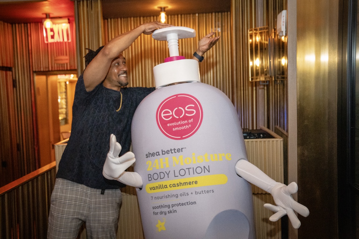 DJ Takeova posing beside the Eos 24H Moisture body lotion mascot at an Eos brand event