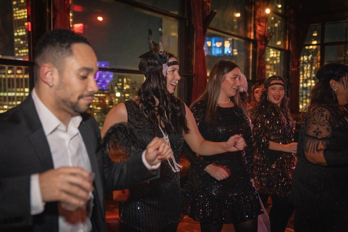Guests in 1920s flapper dresses dancing at a rooftop theme party with the NYC skyline behind them