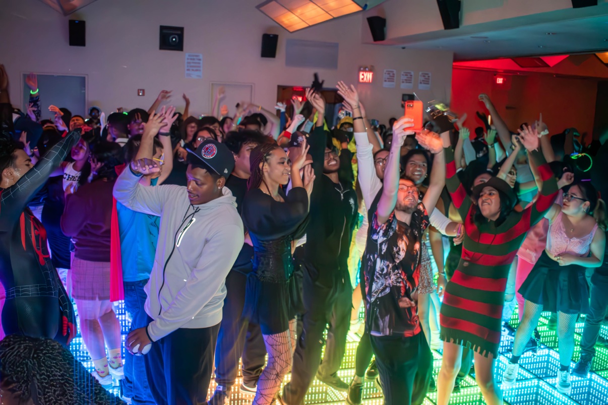 Students in Halloween costumes, including Spider-Man and a Freddy Krueger sweater, dancing with their hands raised on a glowing LED dance floor