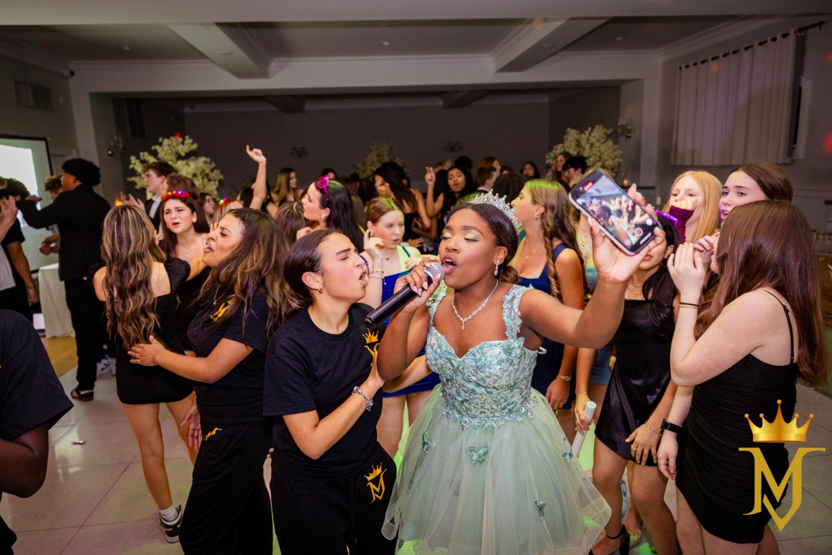 The guest of honor in a mint gown and tiara singing into a microphone, surrounded by friends at her sweet 16