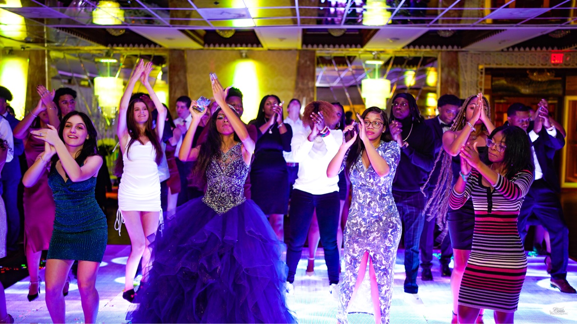 Students in formal wear dancing together on a lit ballroom floor, the guest of honor in a purple gown at the center