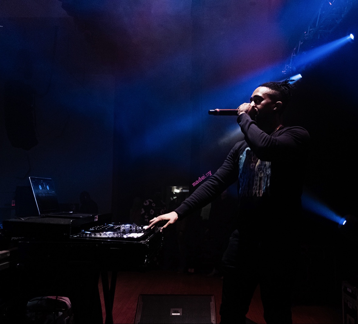 DJ Takeova performing on a concert stage, microphone in hand under blue stage lights