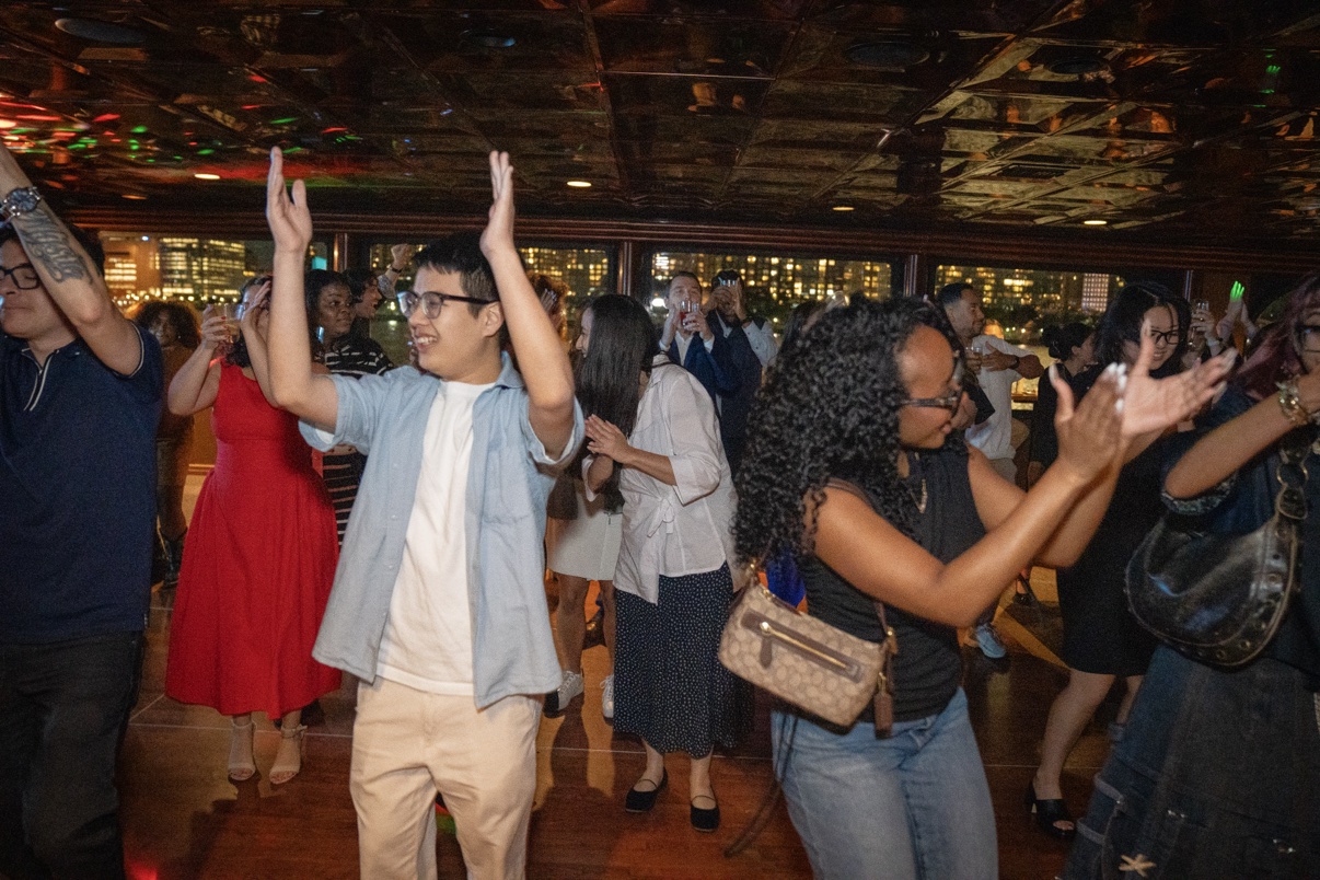 Guests dancing with their hands up at a nighttime birthday boat party with the city skyline visible through the windows