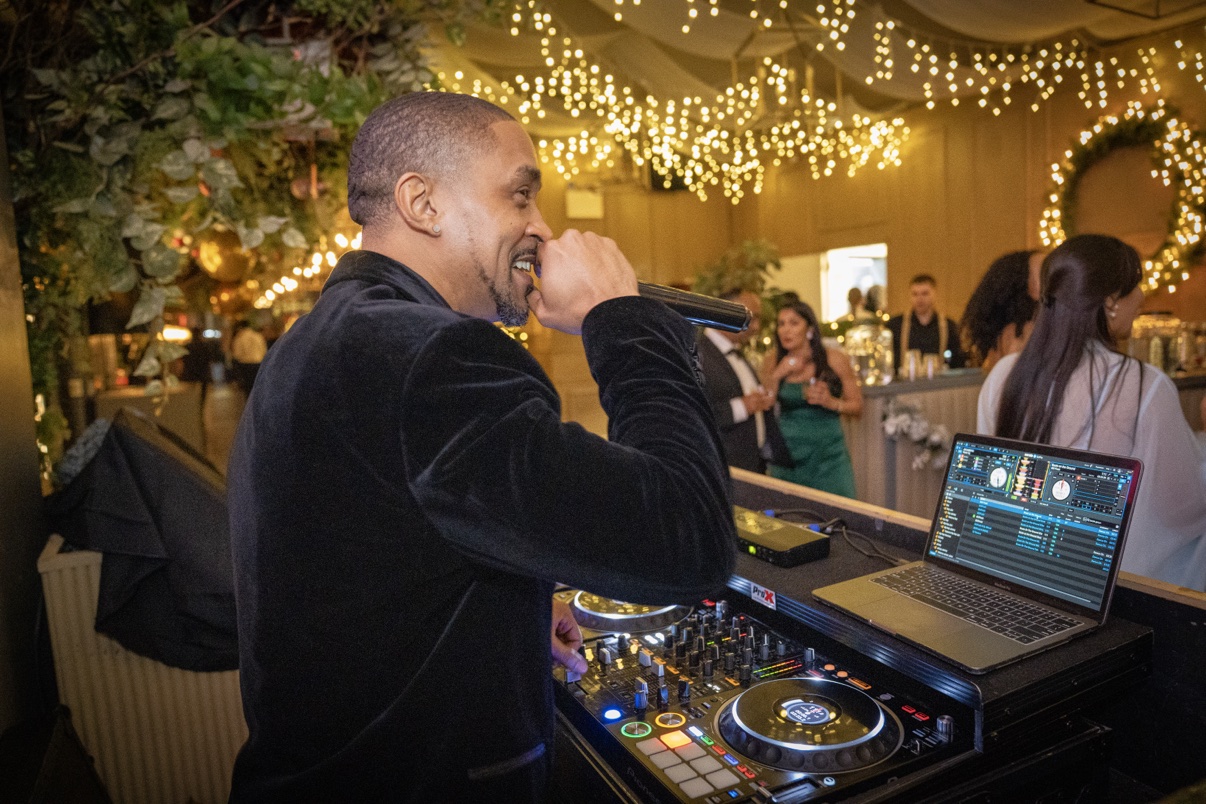 DJ Takeova MCing at the decks in a tuxedo beneath warm string lights at a wedding